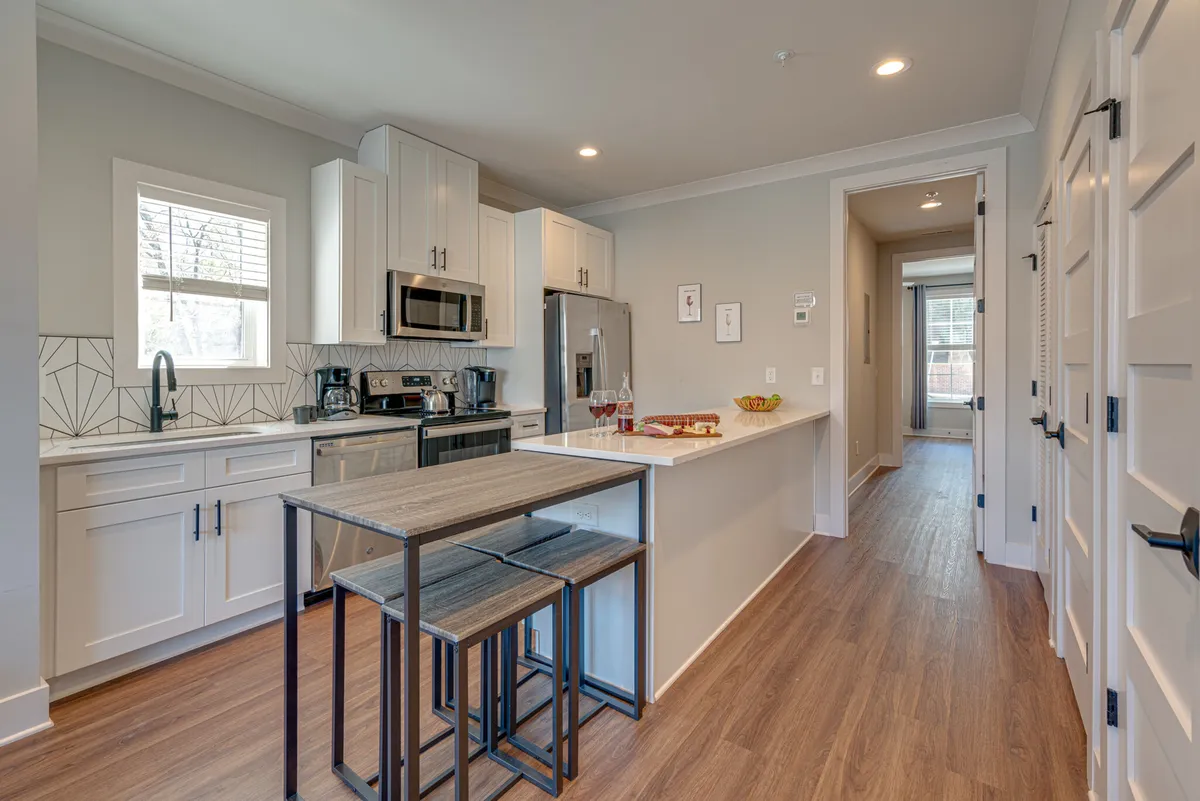 A modern kitchen with stainless steel appliances, an island countertop, and stylish bar stools. The kitchen features decorative wall tiles and is set up for both cooking and entertaining, with plenty of workspace and seating.