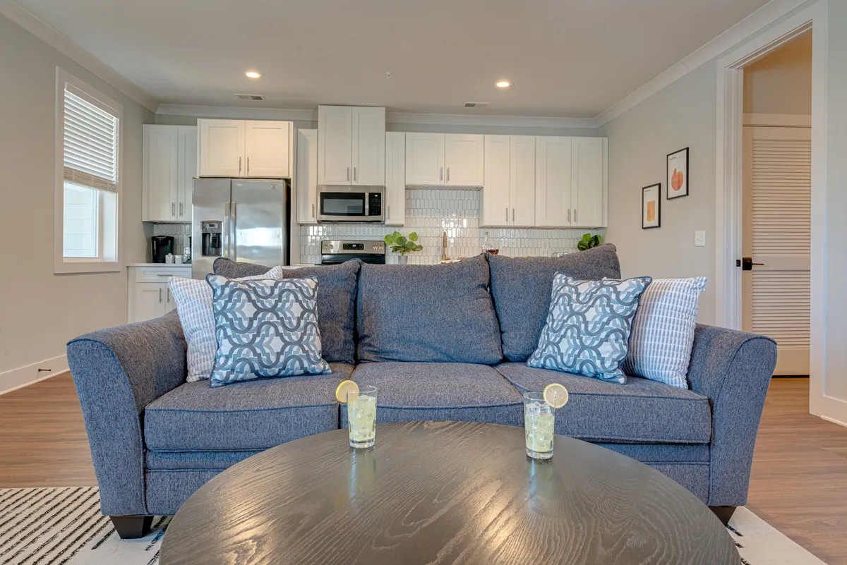 A thoughtfully designed living space featuring a cozy sofa, decorative pillows, a central coffee table, and an open kitchen in the background. The decor is modern yet homey, ideal for both hosting and quiet moments.
