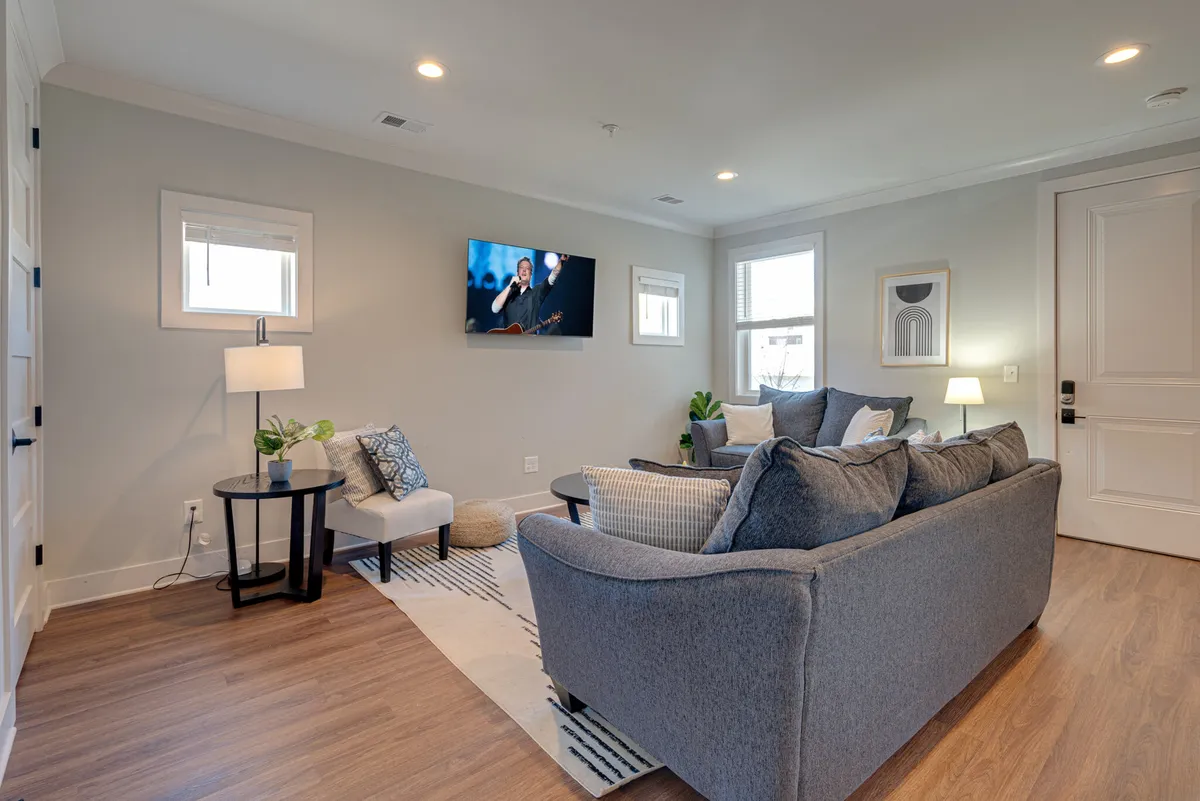 A cozy living room is depicted with a soft sofa, stylish armchair, warm lamp lighting, and a flat-screen television as the focal point. Artwork and thoughtful decor create an inviting atmosphere for movie nights or group downtime.