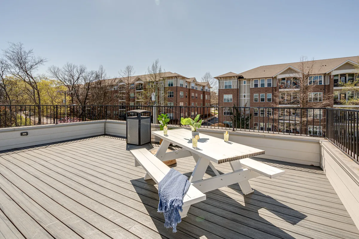 A cheerful rooftop deck scene with a picnic table, green plants, a blue throw blanket, and a trash can. The setup encourages outdoor dining, informal gatherings, and relaxed socializing, with a welcoming ambiance under the sky.