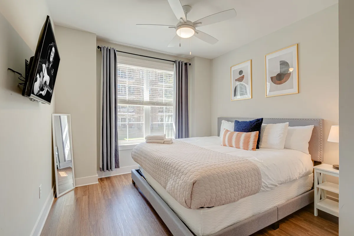 A bright, airy bedroom with a bed adorned with decorative pillows, sunlight streaming through a window, tasteful artwork on the walls, and a stylish mirror. The room exudes a sense of comfort, relaxation, and privacy—ideal for restful nights.