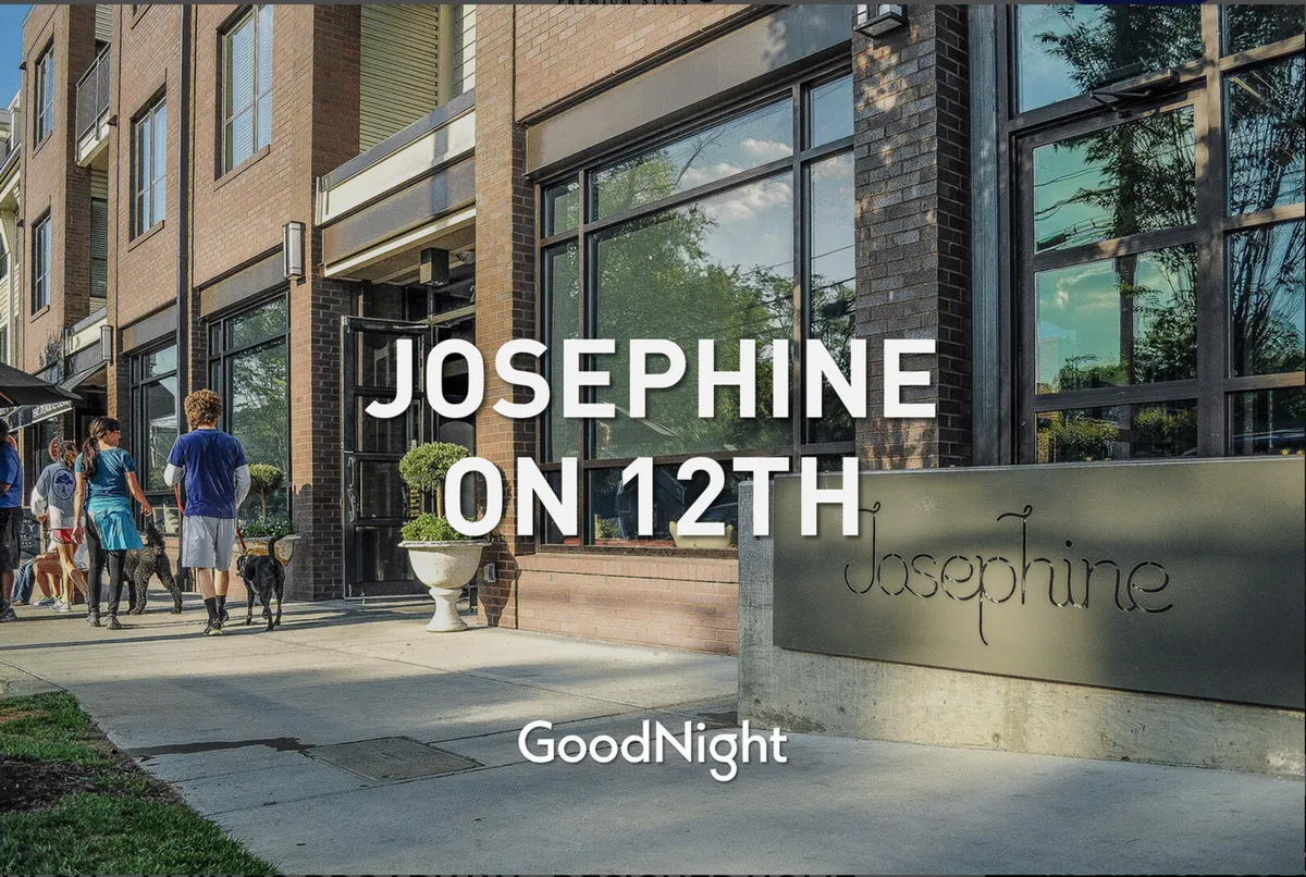 An exterior shot of a building facade featuring clear signage, well-maintained landscaping, and people walking and gathering outside. The scene is active, with individuals walking dogs and socializing, suggesting a friendly, walkable neighborhood atmosphere.