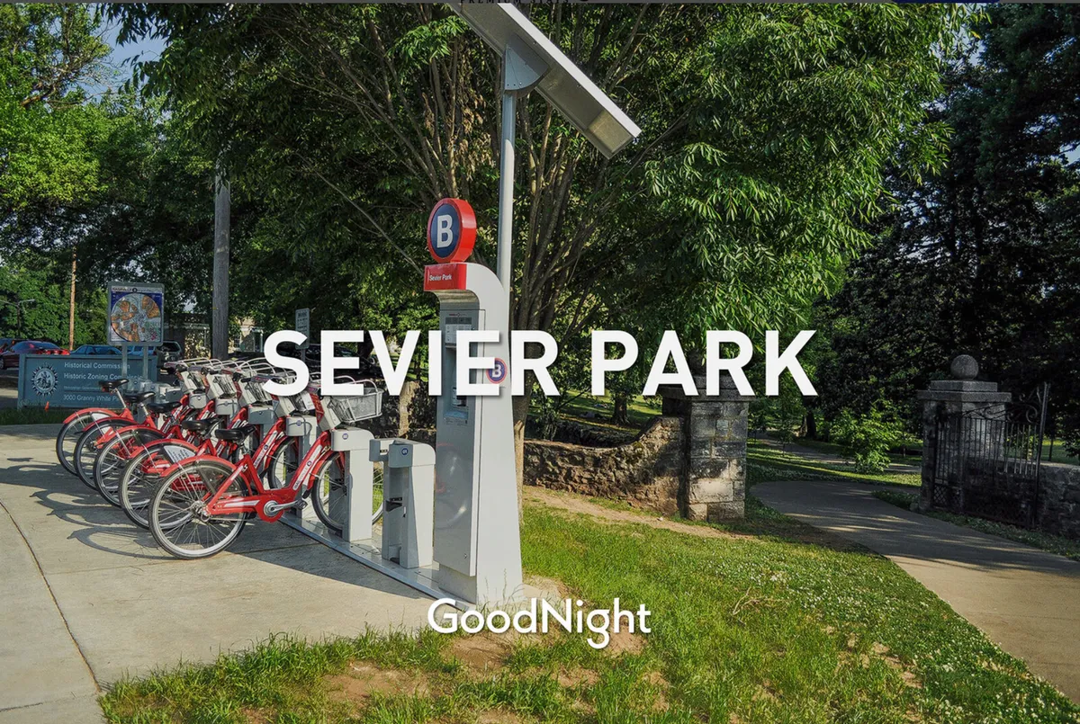 An exterior view featuring a bike-sharing station near a park sign, with lush greenery and a welcoming pathway. The scene suggests easy access to recreational activities for families, such as biking and walking in a nearby park.