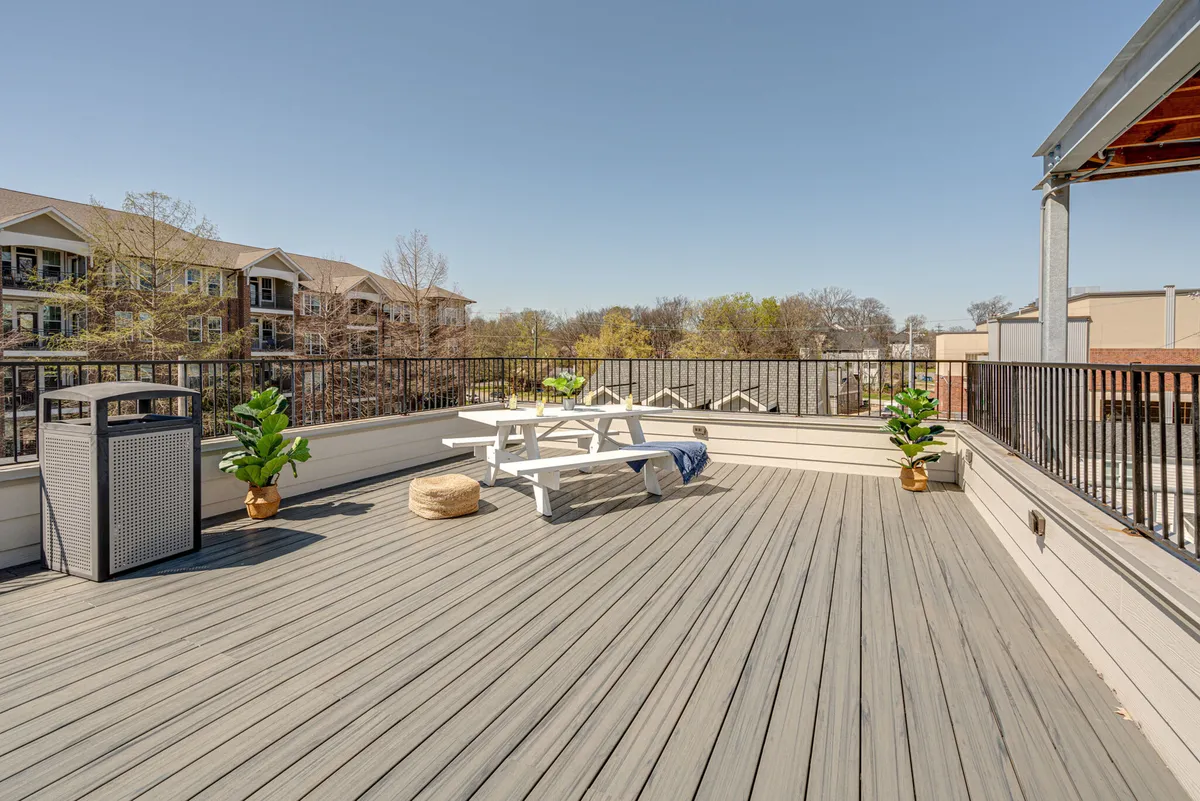 An outdoor rooftop deck featuring a wooden picnic table, surrounded by green plants and a nearby trash can. The deck surface is visible, and the setting appears relaxed, ideal for gatherings or casual meals. The open-air layout suggests space for social activities while offering potential for sunset or skyline views.