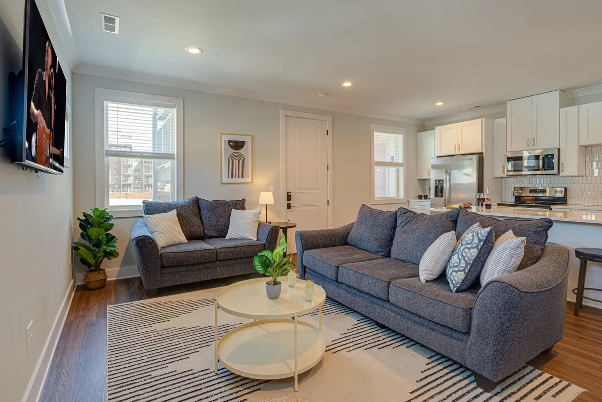 This image features a spacious and inviting living area with comfortable sofas, a central coffee table, vibrant artwork on the wall, and an open view into the kitchen area. The arrangement encourages both relaxation and social interaction, reflecting a multi-functional lounge space ideal for gatherings.