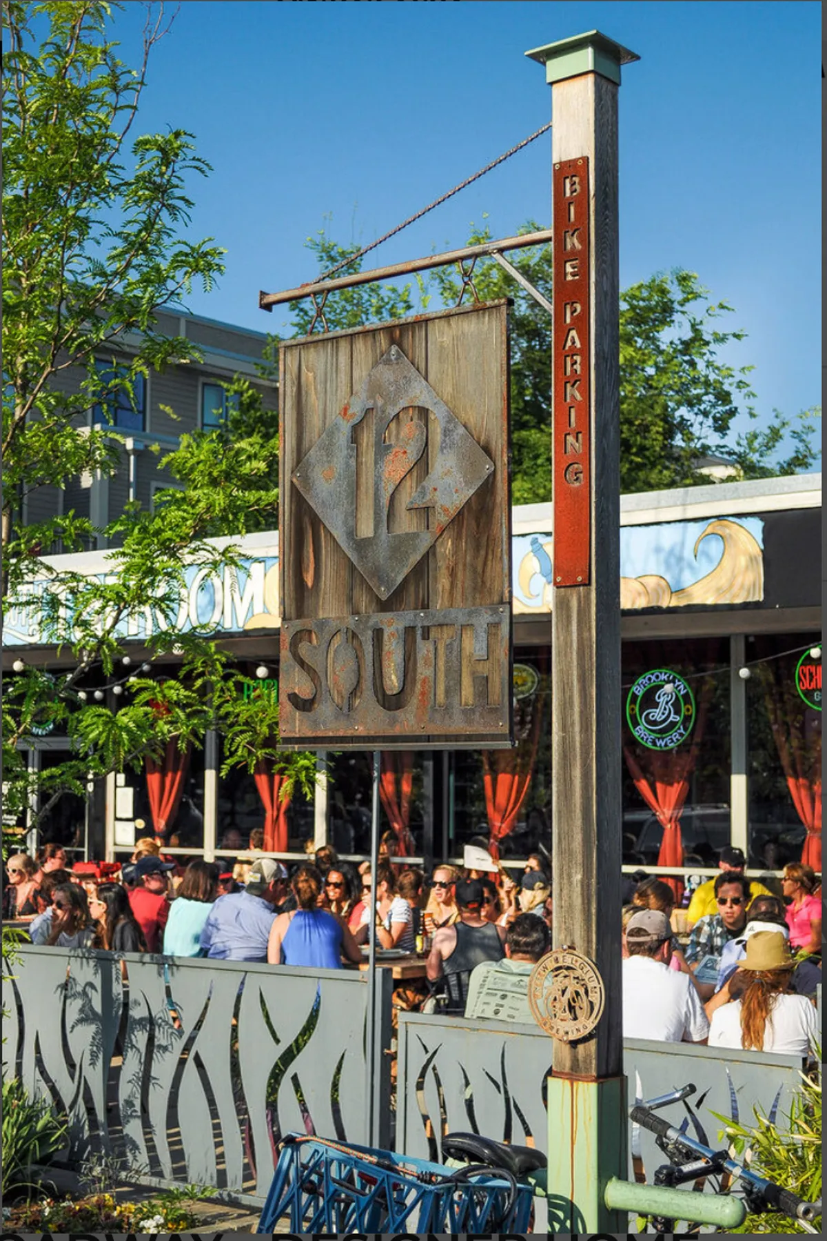 A vibrant exterior scene of a lively neighborhood area with '12 SOUTH' signage, bike parking, and outdoor seating. People are seen enjoying the space, dining, and socializing in a relaxed, community-oriented setting.
