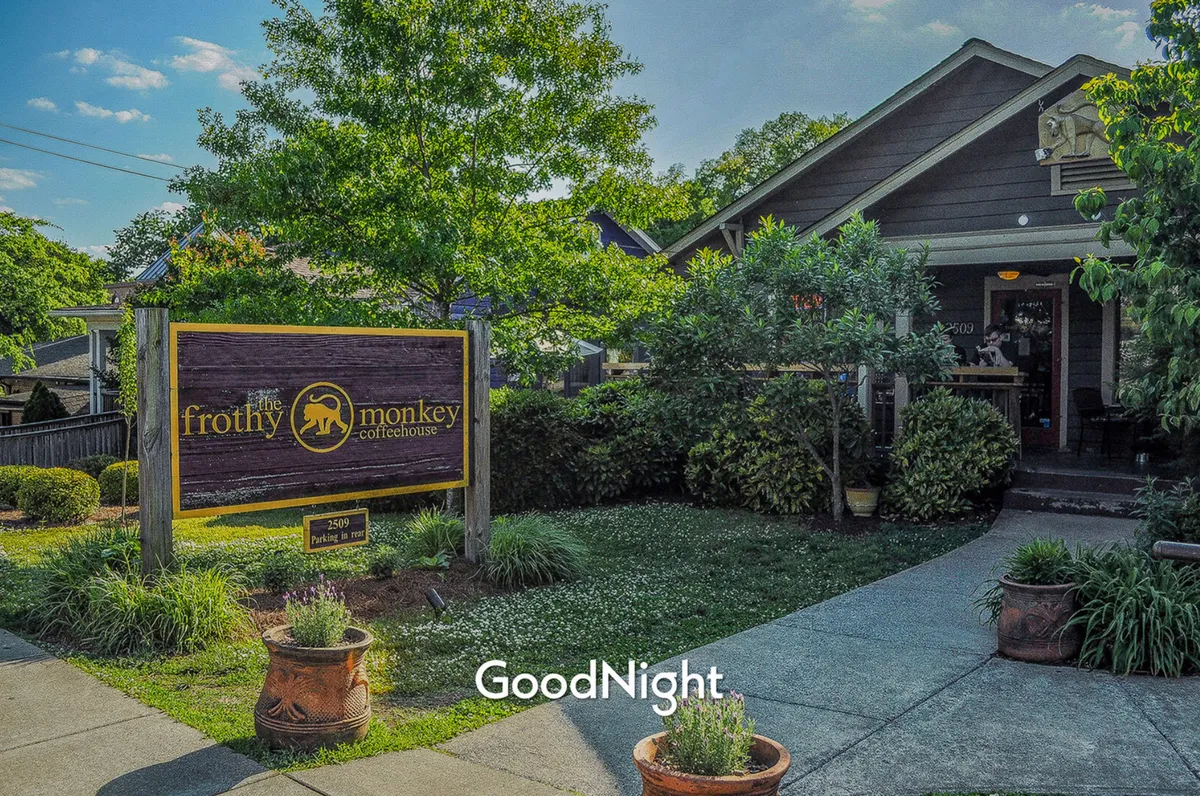 The welcoming entrance of 'The Frothy Monkey' coffeehouse, surrounded by lush landscaping and an inviting outdoor space. The iconic sign and approachable vibe make it a favorite local meeting spot.